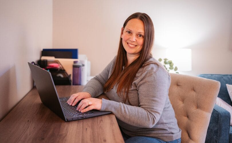 Smiling woman with long brown hair typing on a laptop at a wooden desk in a cozy home office.