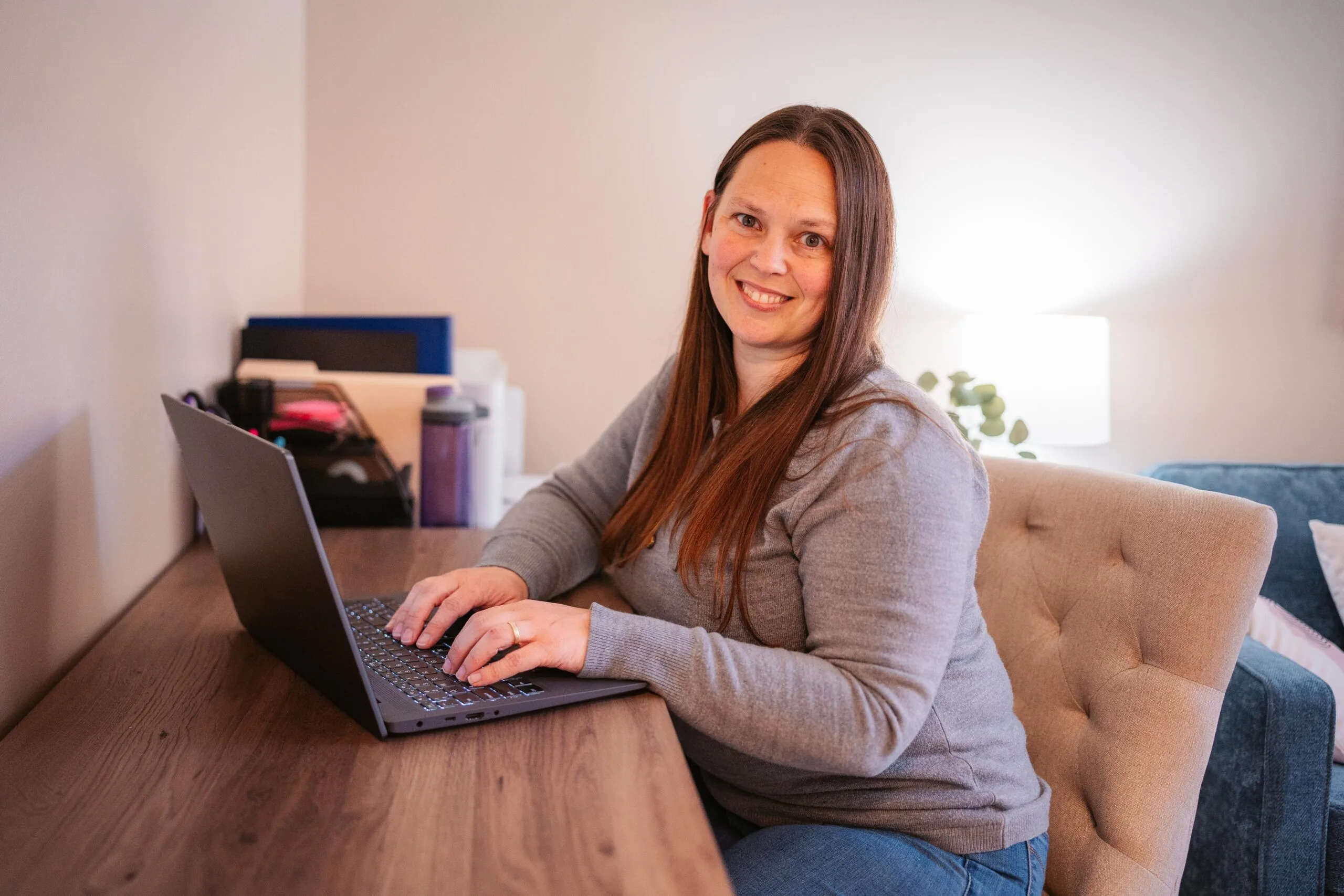 Smiling woman with long brown hair typing on a laptop at a wooden desk in a cozy home office.