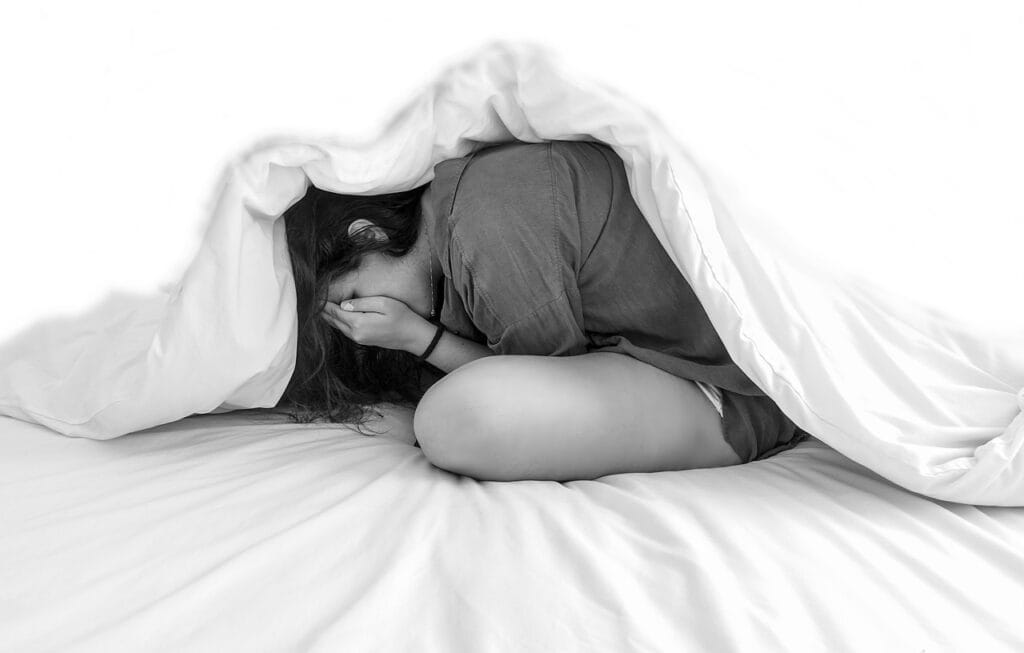 A young woman sits curled up on a bed under a blanket, with her face buried in her hands, expressing sadness or emotional distress.