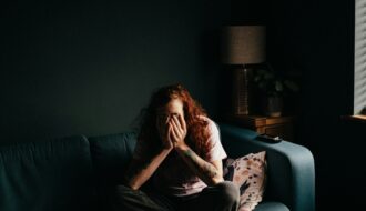Person sitting curled on a couch in a dim room, covering their face with hands, appearing overwhelmed and anxious.