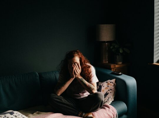 Person sitting curled on a couch in a dim room, covering their face with hands, appearing overwhelmed and anxious.