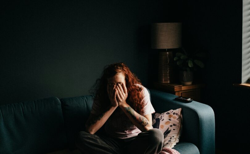 Person sitting curled on a couch in a dim room, covering their face with hands, appearing overwhelmed and anxious.