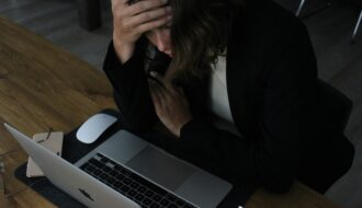 A therapist sits at a desk with her head in her hands, visibly overwhelmed, staring down at an open laptop.