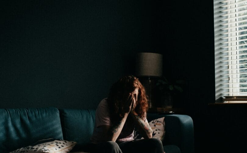 A woman sitting alone on a couch in a dimly lit room with her face in her hands