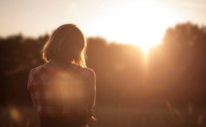 woman standing alone in a field facing a warm golden sunset