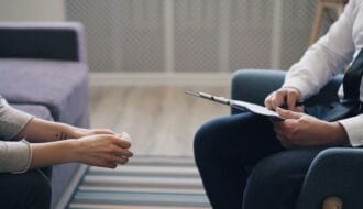 A therapist holding a clipboard listens to a client during a grief therapy session for chronic illness.