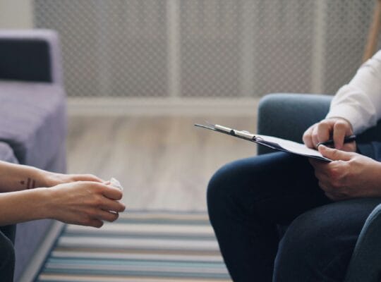 A therapist holding a clipboard listens to a client during a grief therapy session for chronic illness.