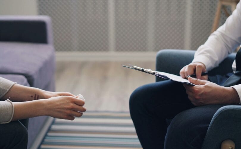 A therapist holding a clipboard listens to a client during a grief therapy session for chronic illness.