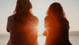 Two women sit together watching the sunset over the ocean, representing companionship through anticipatory grief