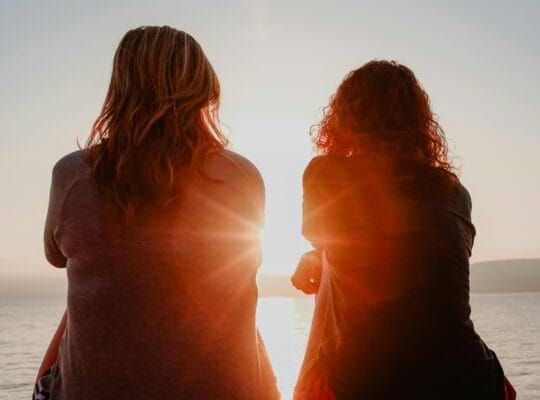 Two women sit together watching the sunset over the ocean, representing companionship through anticipatory grief