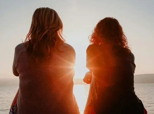 Two women sit together watching the sunset over the ocean, representing companionship through anticipatory grief