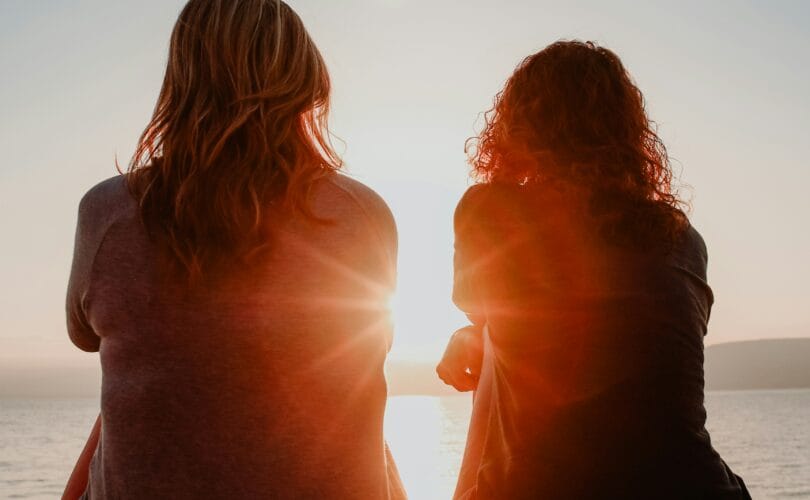Two women sit together watching the sunset over the ocean, representing companionship through anticipatory grief