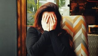 Woman sitting in a chair with her face buried in her hands, wearing a black sweater, conveying emotional exhaustion and overwhelm.