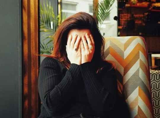 Woman sitting in a chair with her face buried in her hands, wearing a black sweater, conveying emotional exhaustion and overwhelm.