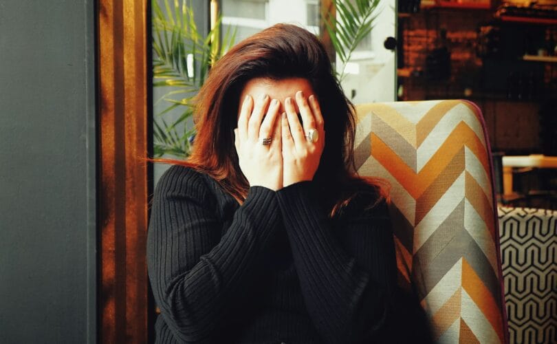 Woman sitting in a chair with her face buried in her hands, wearing a black sweater, conveying emotional exhaustion and overwhelm.