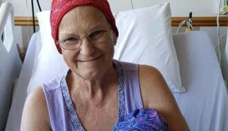Woman in a hospital bed wearing a red headscarf, smiling while holding a stuffed Eeyore wearing a matching blue headscarf