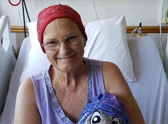 Woman in a hospital bed wearing a red headscarf, smiling while holding a stuffed Eeyore wearing a matching blue headscarf