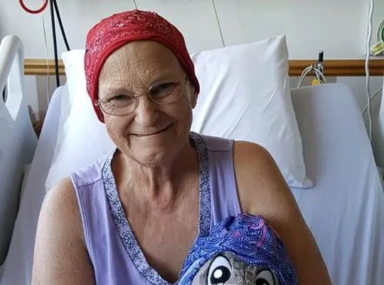 Woman in a hospital bed wearing a red headscarf, smiling while holding a stuffed Eeyore wearing a matching blue headscarf