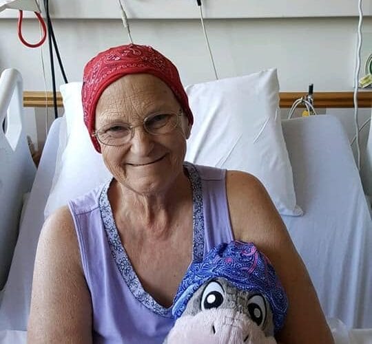 Woman in a hospital bed wearing a red headscarf, smiling while holding a stuffed Eeyore wearing a matching blue headscarf