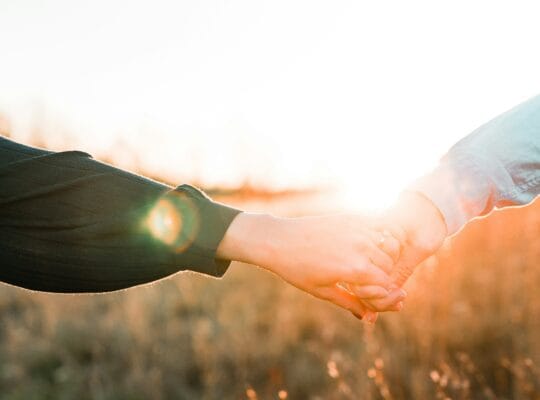 Two people holding hands outdoors at sunset, sunlight glowing through a golden field