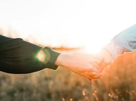 Two people holding hands outdoors at sunset, sunlight glowing through a golden field