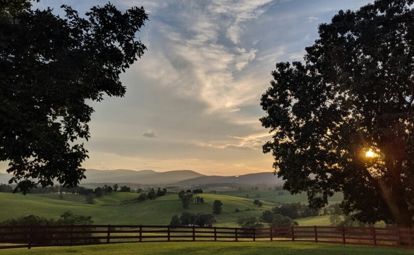 Sunset over rolling green hills and farmland in rural Virginia, framed by two large trees and a wooden fence