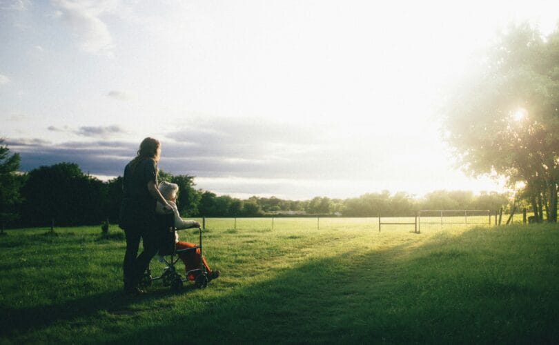 Woman pushing an elderly loved one in a wheelchair through a field at sunset
